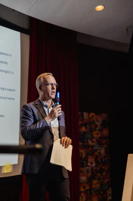 A close shot of the presenter mid-talk, mic in one hand and notes dangling in the other, speaking with an open, animated expression. A partial projected slide is visible to the left and the curved ceiling of the auditorium appears above.