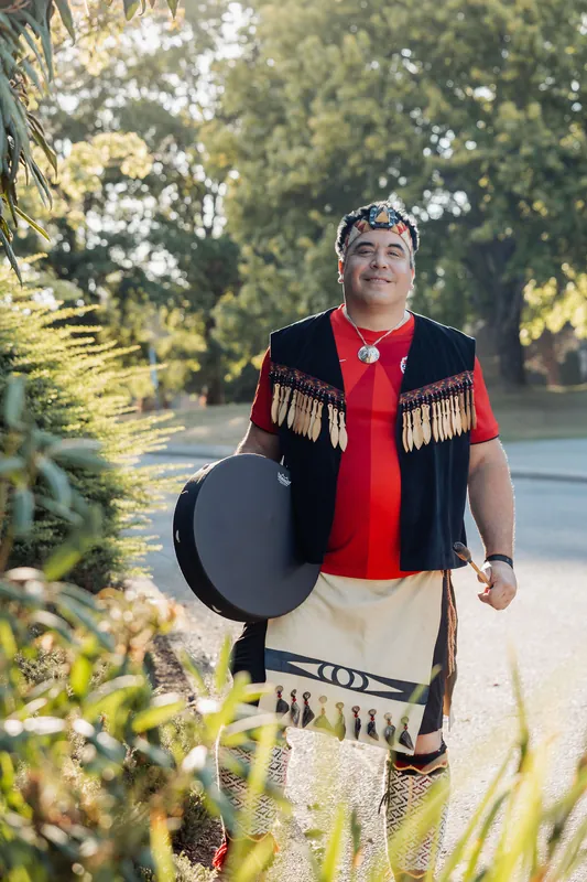 A performer in traditional fringed regalia and headband walks the sun-dappled grounds carrying a large black hand drum.