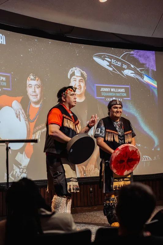 Two performers in traditional regalia stand side by side on stage as their Squamish Nation titles appear on the projection screen.