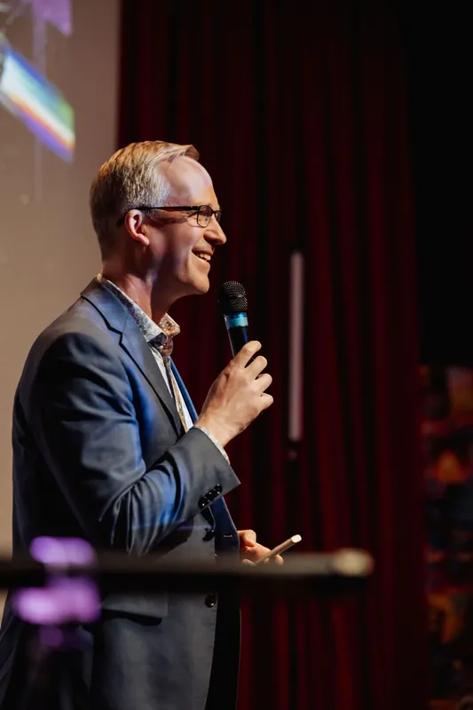 A laughing emcee holds court on stage, presentation slide glowing behind him.