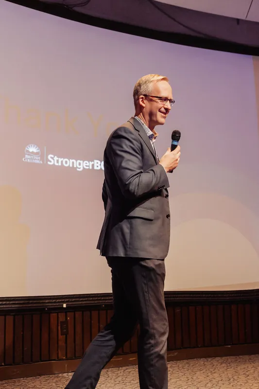 The speaker stands on the stage floor with a wide smile and relaxed posture, mic held loosely at their side. A branded slide glows warm on the curved projection screen behind them.