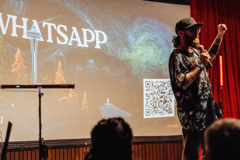 A presenter in a baseball cap pumps a fist toward the ceiling as a WhatsApp community-join slide glows behind him on stage.