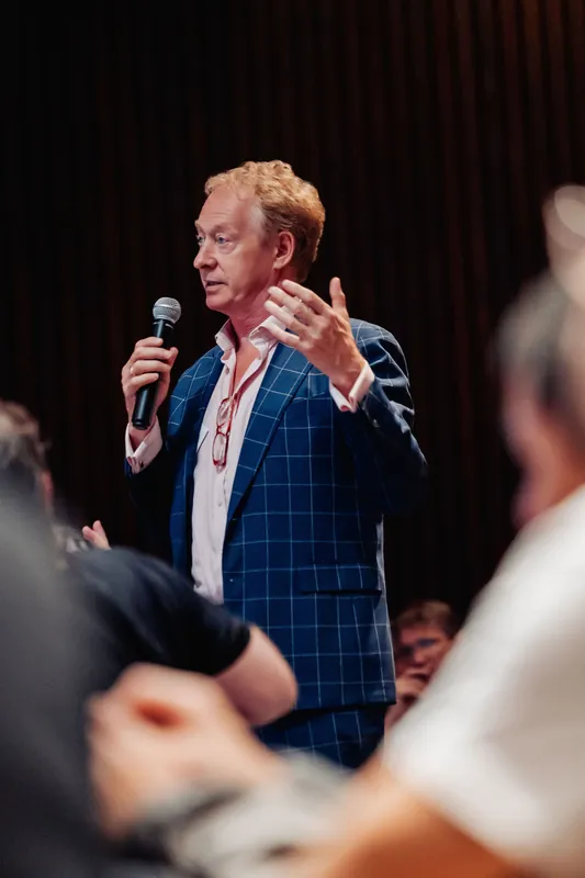 A speaker holds court mid-sentence as a close foreground of audience members frames the shot.