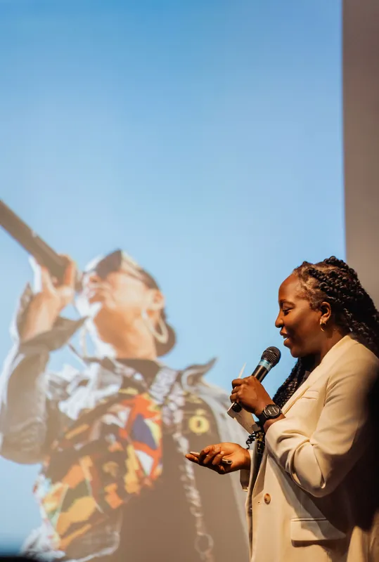 Anthonia Ogundele holds the mic as a vivid performer image fills the blue projection screen behind her.