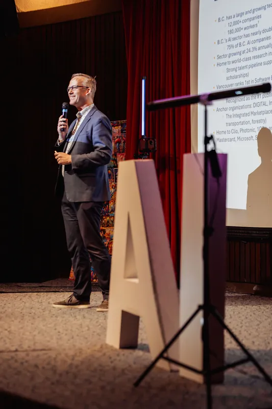 A speaker presents BC's AI sector highlights from the stage, a large letter A prop and projection screen framing the moment.