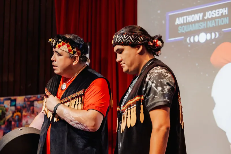 The two Squamish Nation performers stand ready at the front of the stage, their names displayed in the projection overhead.