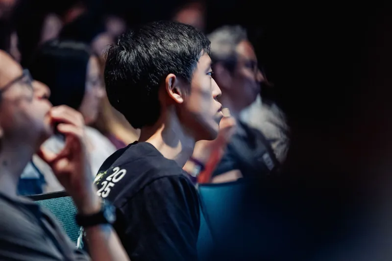 A young attendee in a black tee watches the stage with focused intensity, his face caught in a slant of warm stage light.