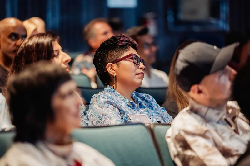 A woman in red-framed glasses looks attentively toward the stage, surrounded by a full row of engaged audience members.
