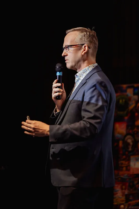Framed from slightly below and behind, the presenter holds the mic close to their lips and extends the free hand forward with relaxed, open fingers. A vivid mural or display fills the background at right.