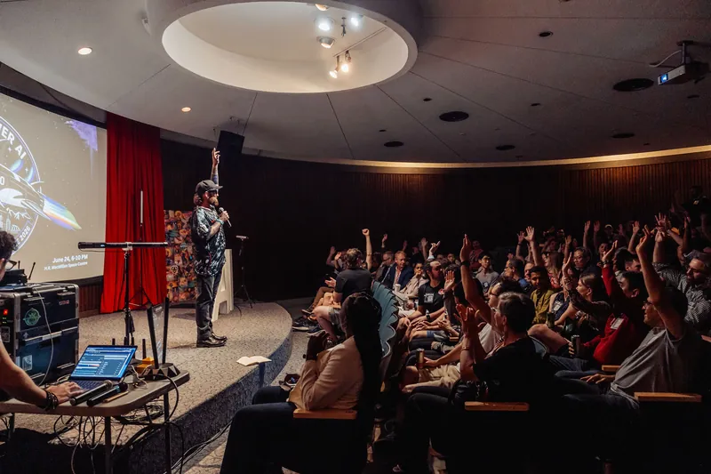 A presenter raises a fist at the mic as dozens of hands shoot up across the packed planetarium theatre.