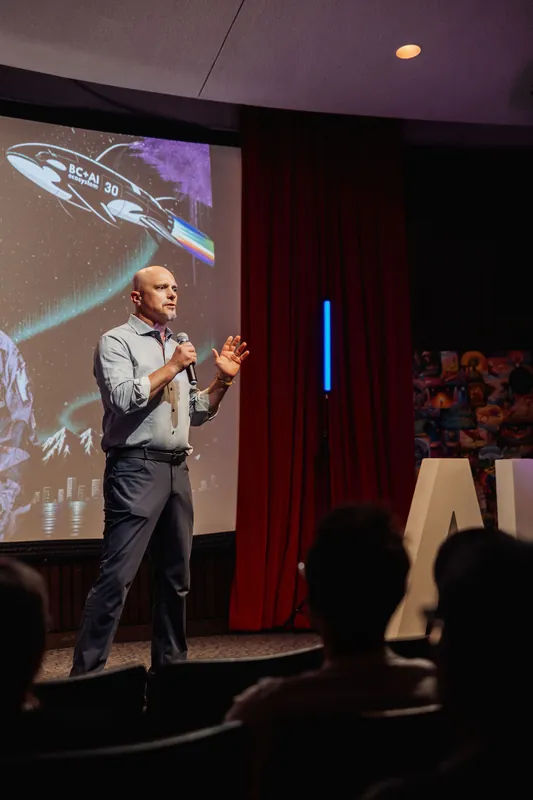 A speaker gestures mid-thought while presenting to a seated crowd beneath the BC+AI 30 starfield projection.