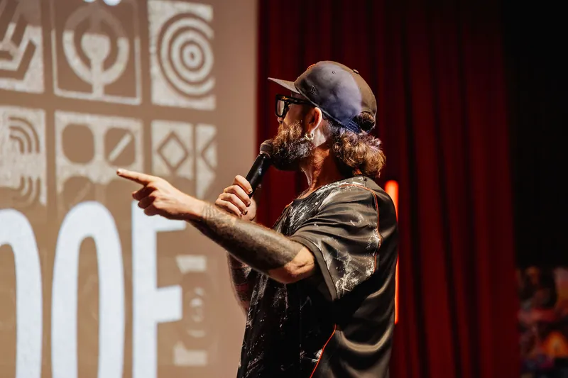 A presenter points emphatically toward the crowd, geometric and tribal-pattern projection filling the wall behind him.