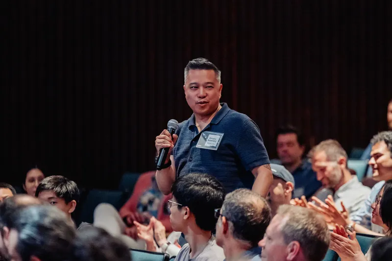 An attendee stands with a mic as the packed crowd around him applauds warmly.