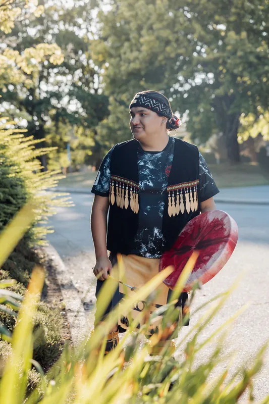 Bathed in warm afternoon sun, a Squamish Nation performer strolls a garden path holding a red ceremonial drum.