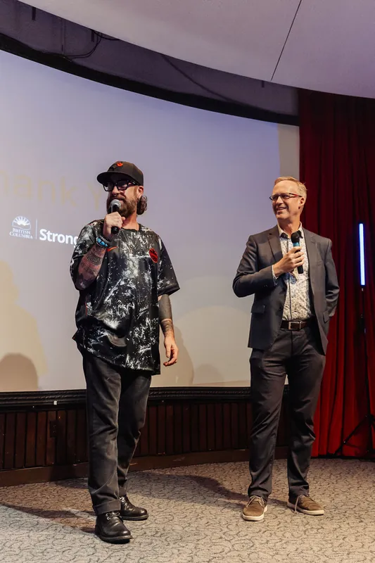 Two presenters share the stage — one in a baseball cap and graphic tee, the other in a blazer — both holding mics and facing the audience in what appears to be a warm handoff between segments.