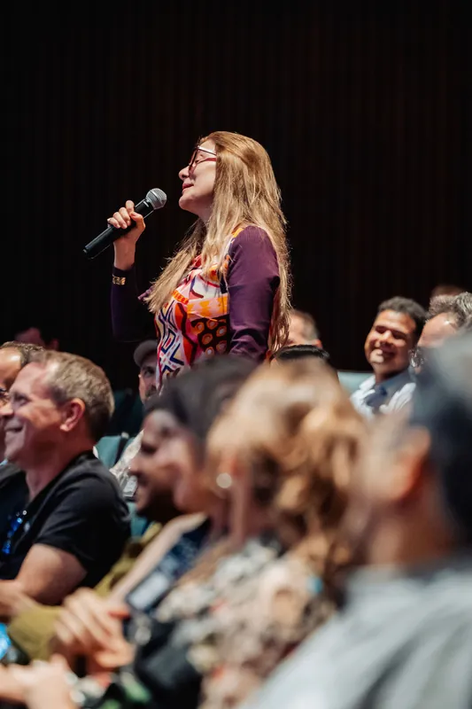 A woman with long reddish hair speaks into the mic, her colorful patterned outfit vivid against the surrounding crowd.