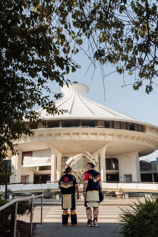 Two Indigenous performers in traditional regalia approach the H.R. MacMillan Space Centre, framed by summer foliage in golden light.