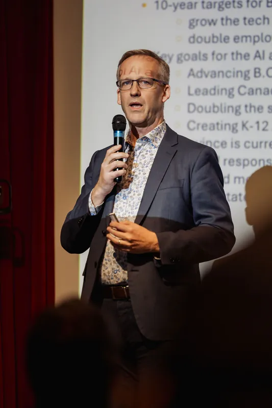 The speaker stands upright with the mic raised to speaking height, mouth open mid-delivery. A lit projection screen behind them shows a bulleted slide. Two silhouetted heads are visible in the lower corners of the frame.