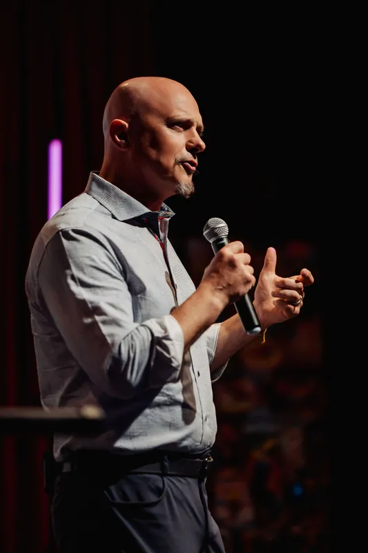 Close-up side portrait of a presenter mid-sentence, hands moving with emphasis against the dark warm light of the venue.