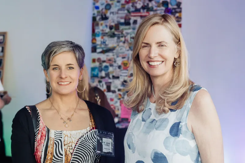 Two guests beam at the camera in front of a colourful collage backdrop during the evening networking reception.