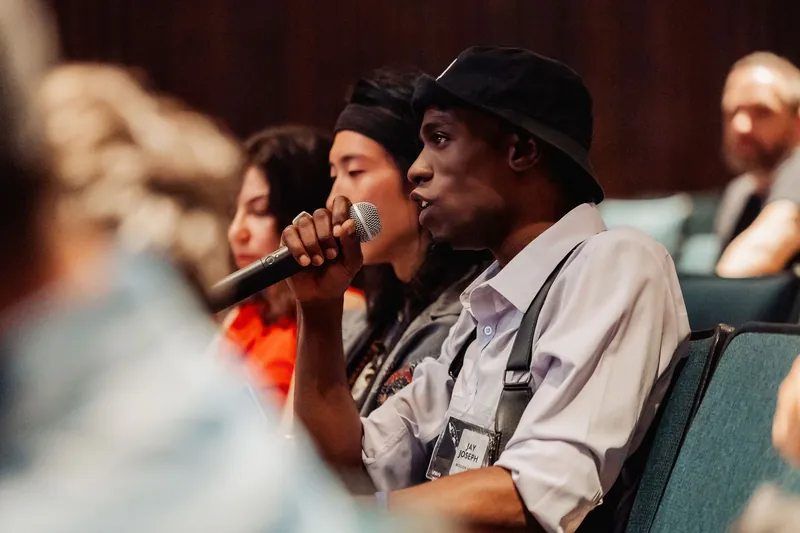A seated audience member leans forward holding a handheld microphone to their mouth, appearing to speak during a Q&A exchange. Several other attendees are visible in soft focus on either side.