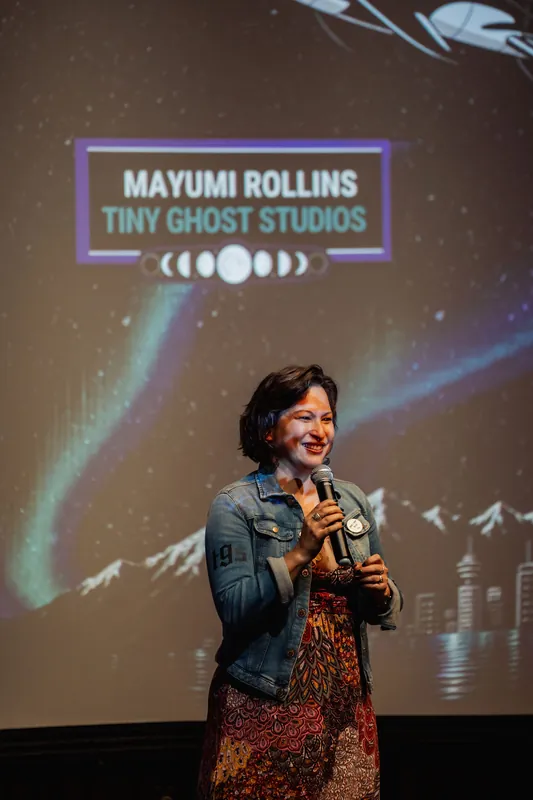 Mayumi Rollins of Tiny Ghost Studios smiles mid-talk on stage beneath a sweeping aurora projection inside the Space Centre.