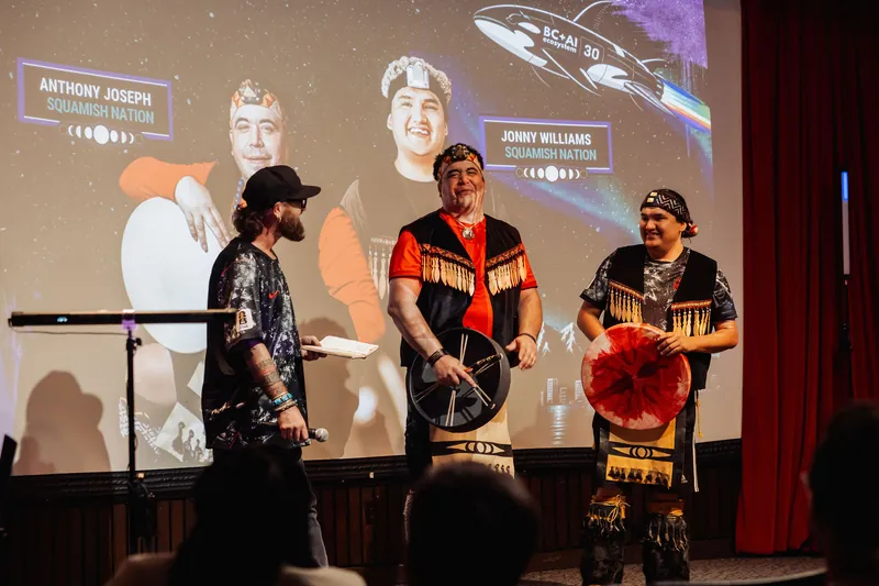 Three on stage: the host and two Squamish Nation performers with drums, smiling warmly for the crowd.