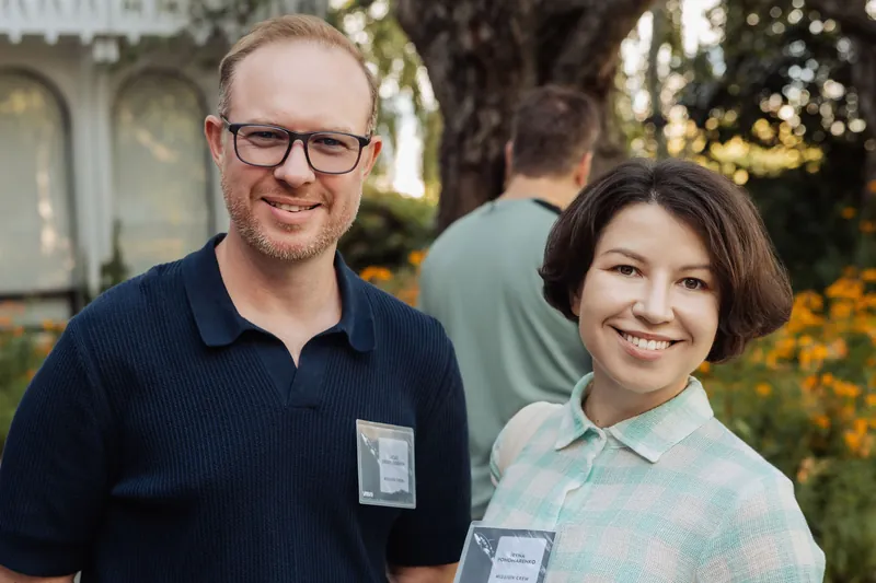 Two attendees smile toward the camera as soft evening light filters through the garden trees behind them.