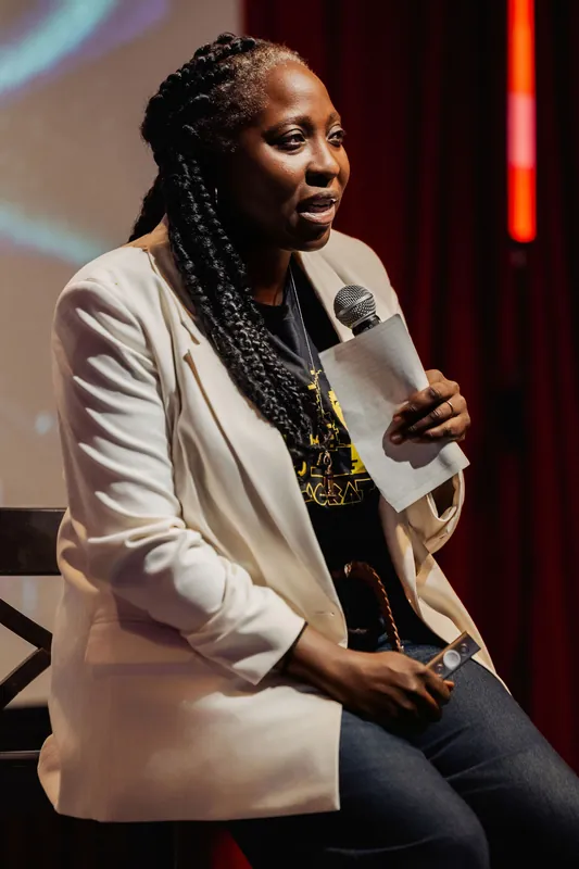 Anthonia Ogundele is captured mid-thought on stage, mic raised and papers in her lap, the red curtain warm behind her.