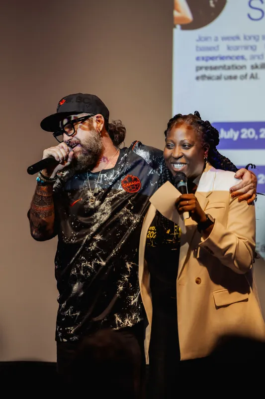 The tattooed host and Anthonia Ogundele share a warm laugh together on stage, both holding microphones.