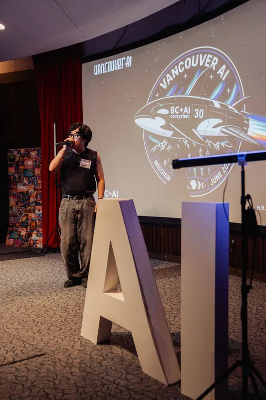 The MC addresses the audience from beside the event's oversized letter-form AI installation, microphone raised.
