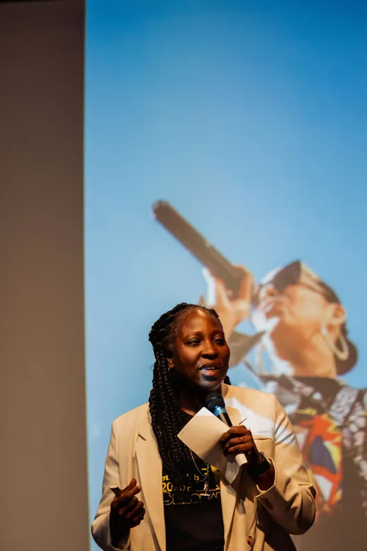 Anthonia Ogundele speaks at the podium while a bold close-up of a performer is projected on the screen.