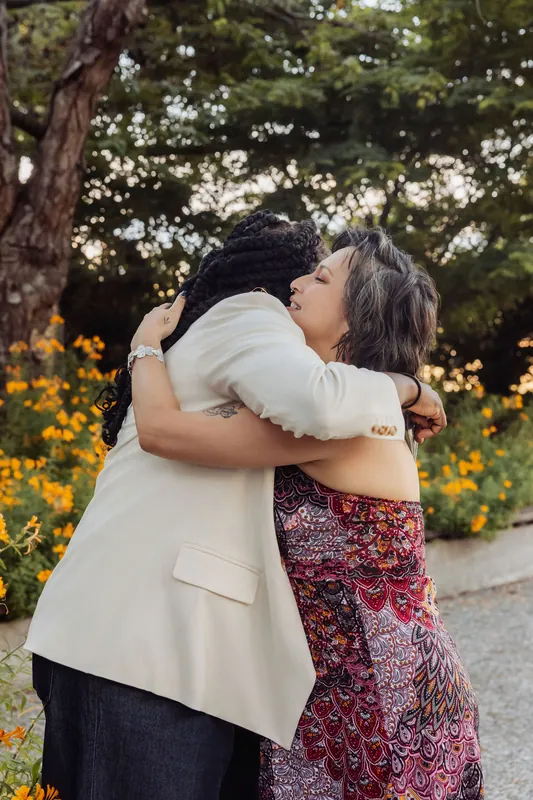 Two women share a heartfelt embrace in the flower garden as golden-hour light glows softly behind them.