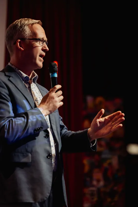 A close-up of the same presenter mid-sentence, mouth open, one hand gripping the microphone and the other raised in a cupped, open gesture at waist height. The red stage curtains and a blurred row of seated attendees are softly visible behind them.