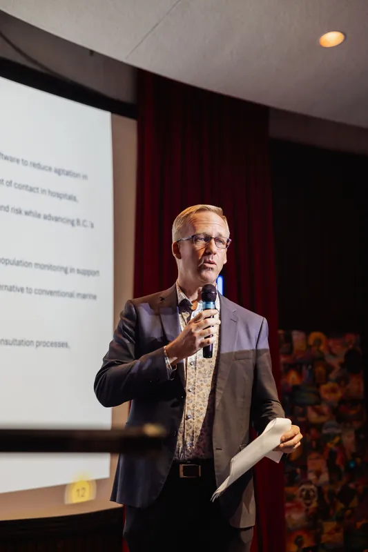 A three-quarter view of the presenter holding a mic and a folded sheet of notes, gazing forward with a composed expression. A projected slide occupies the left of the frame while a colorful tapestry-style artwork hangs on the wall behind the stage at right.