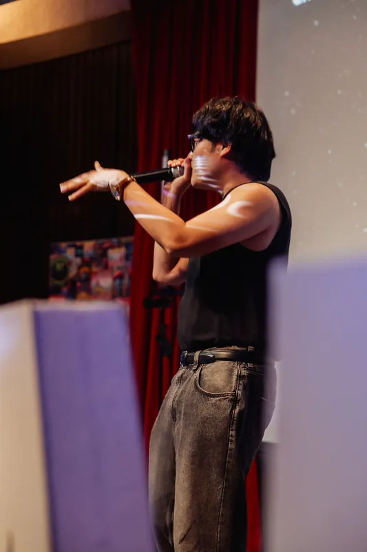 The performer extends an arm in a dramatic mid-set gesture, mic held close in the darkened theater.