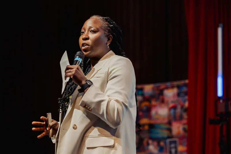 Now standing, Anthonia Ogundele gestures with an open palm while speaking to the full room, colorful artwork glowing behind her.
