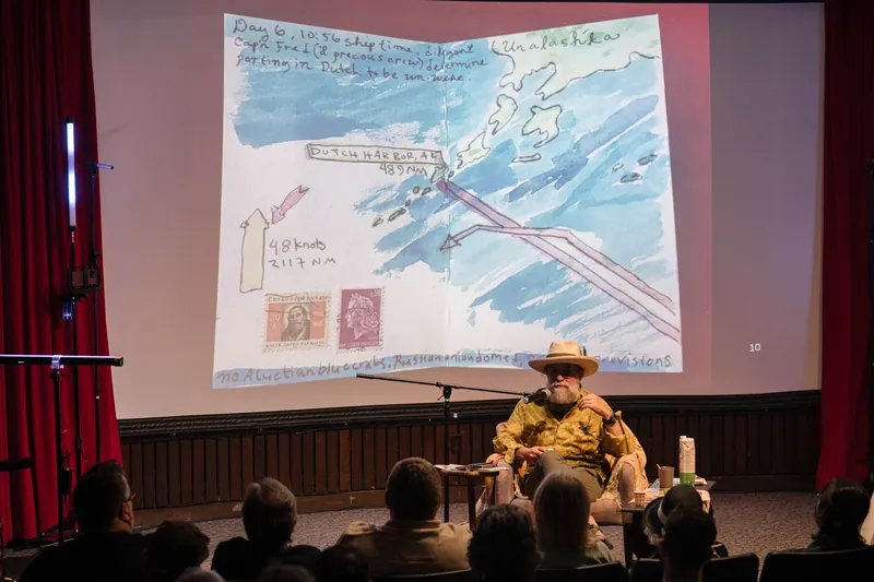Dave Olson speaks from the armchair, gesturing as slides cycle behind him — Screen bright in the background.