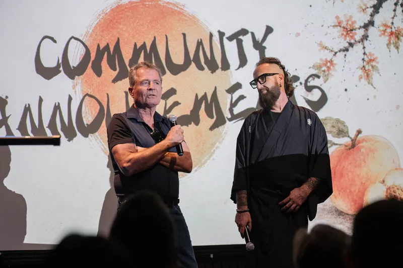 More community announcements roll on — mic handoffs and short stage spots — Audience heads in the foreground.