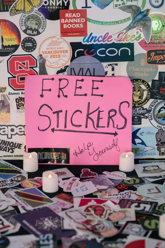 A second angle on the free-stickers table — pink Help Yourself sign, tea lights, and loose Vancouver.ai stickers.