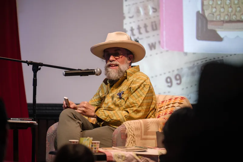 Dave Olson speaks from the armchair, gesturing as slides cycle behind him — From the aisle.