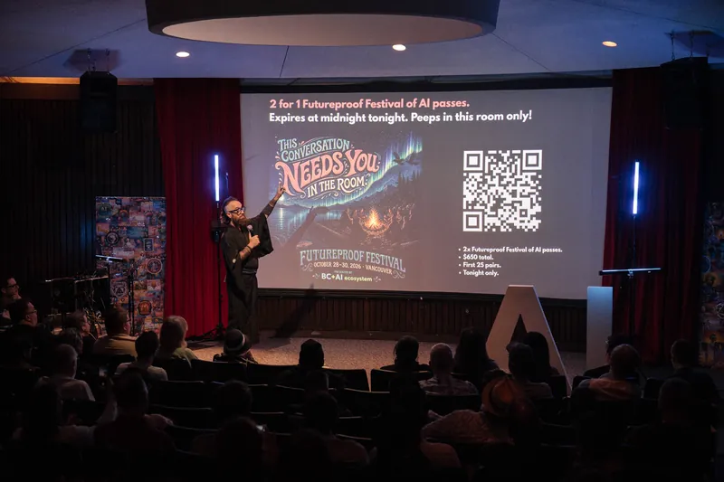 A wide shot from the back of the room — silhouetted audience facing the lit stage and screen.