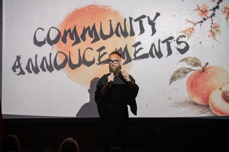 Community Announcements open with Kris on stage beside the peach-framed slide — Stage-left view.