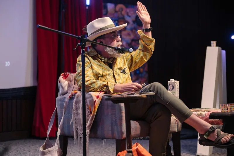 Dave Olson, in hat and yellow shirt, raises a hand mid-story from the stage armchair.