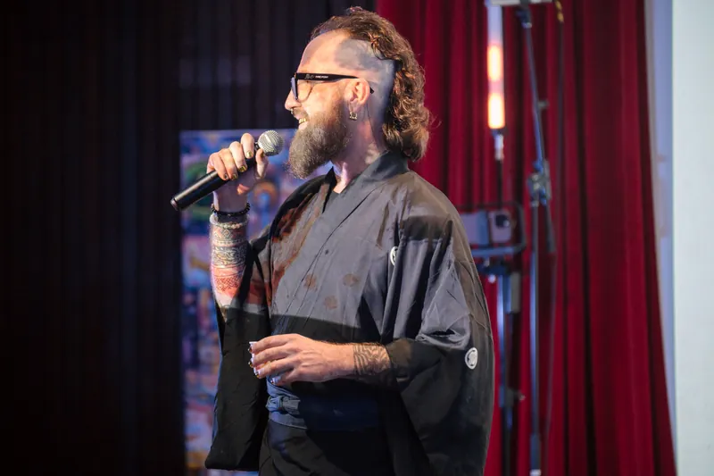 A performer with a mohawk speaks into the mic against the red stage curtain.