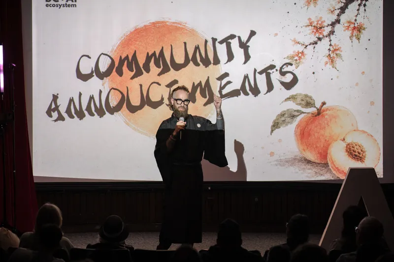 Community Announcements open with Kris on stage beside the peach-framed slide — From the aisle.