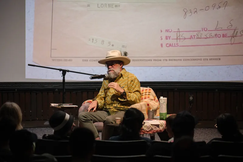 Dave Olson leans into storytelling — clippings, collages, and handwritten notes on screen — Audience heads in the foreground.