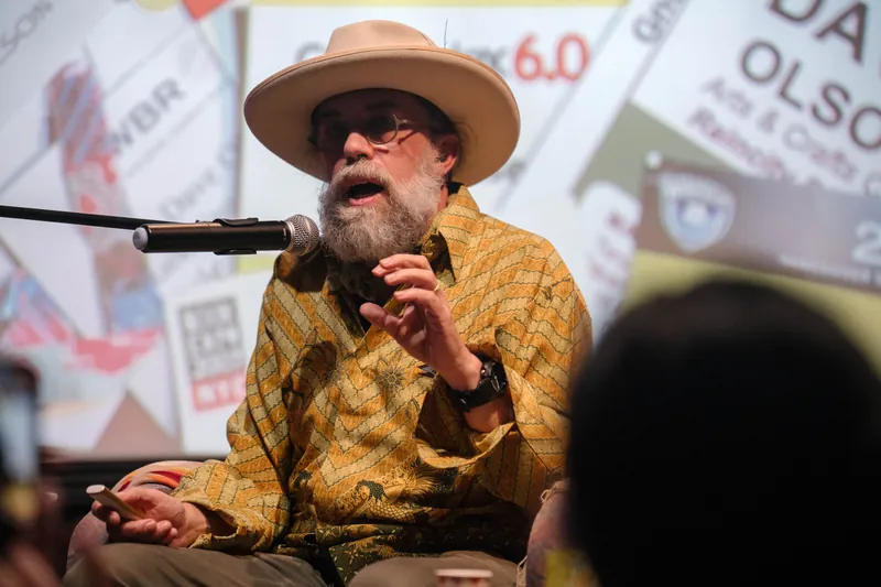 Dave Olson continues from the armchair as denser text and photo slides roll — Soft focus on the crowd edge.