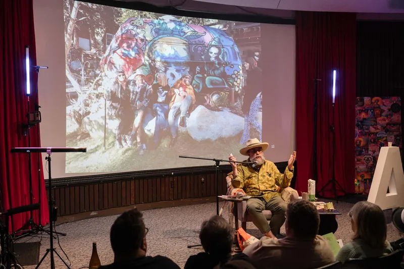 Dave Olson speaks from the armchair, gesturing as slides cycle behind him — A beat later in the same beat.