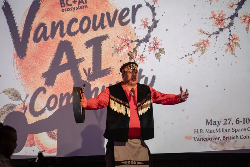 An Indigenous welcome performer holds a hand drum wide on the Vancouver AI Community stage.